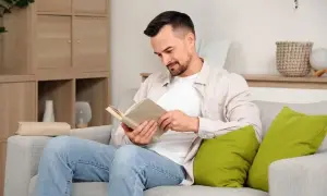 Mężczyzna w jasnej koszuli i jeansach siedzi na kanapie i czyta książkę.