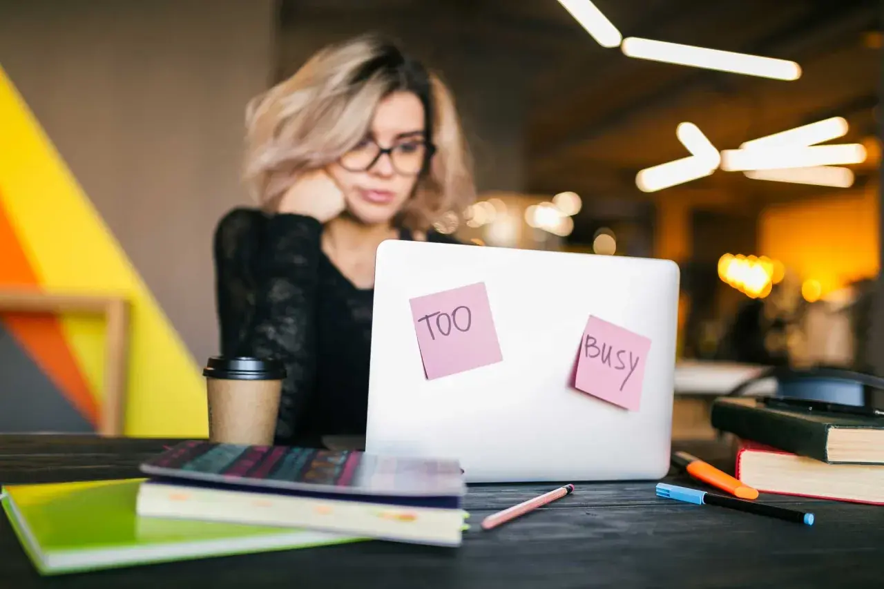 Kobieta z okularami przy laptopie, na którym karteczki "Too busy". Nawet przy natłoku obowiązków, samodzielna nauka angielskiego jest możliwa.