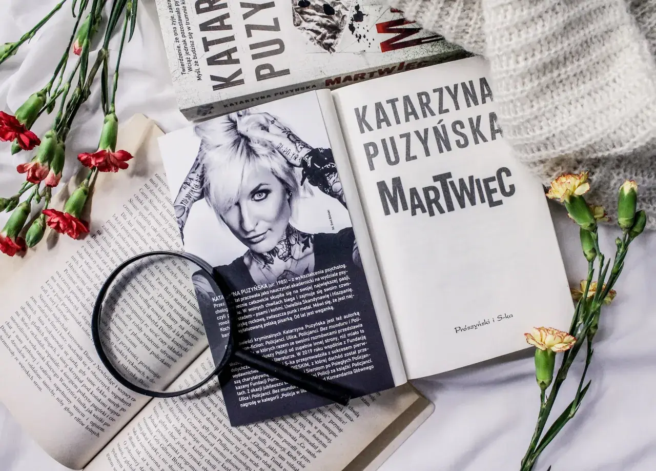 Książki Katarzyny Puzyńskiej, kwiaty i szkło powiększające na białym tle.