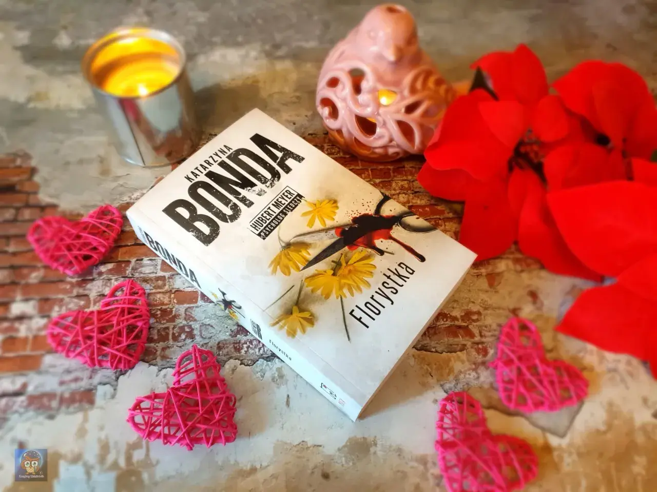 Książka "Florystka" z serii Katarzyny Bondy, z motywem Huberta Meyera, otoczona walentynkowymi serduszkami i płonącą świecą.