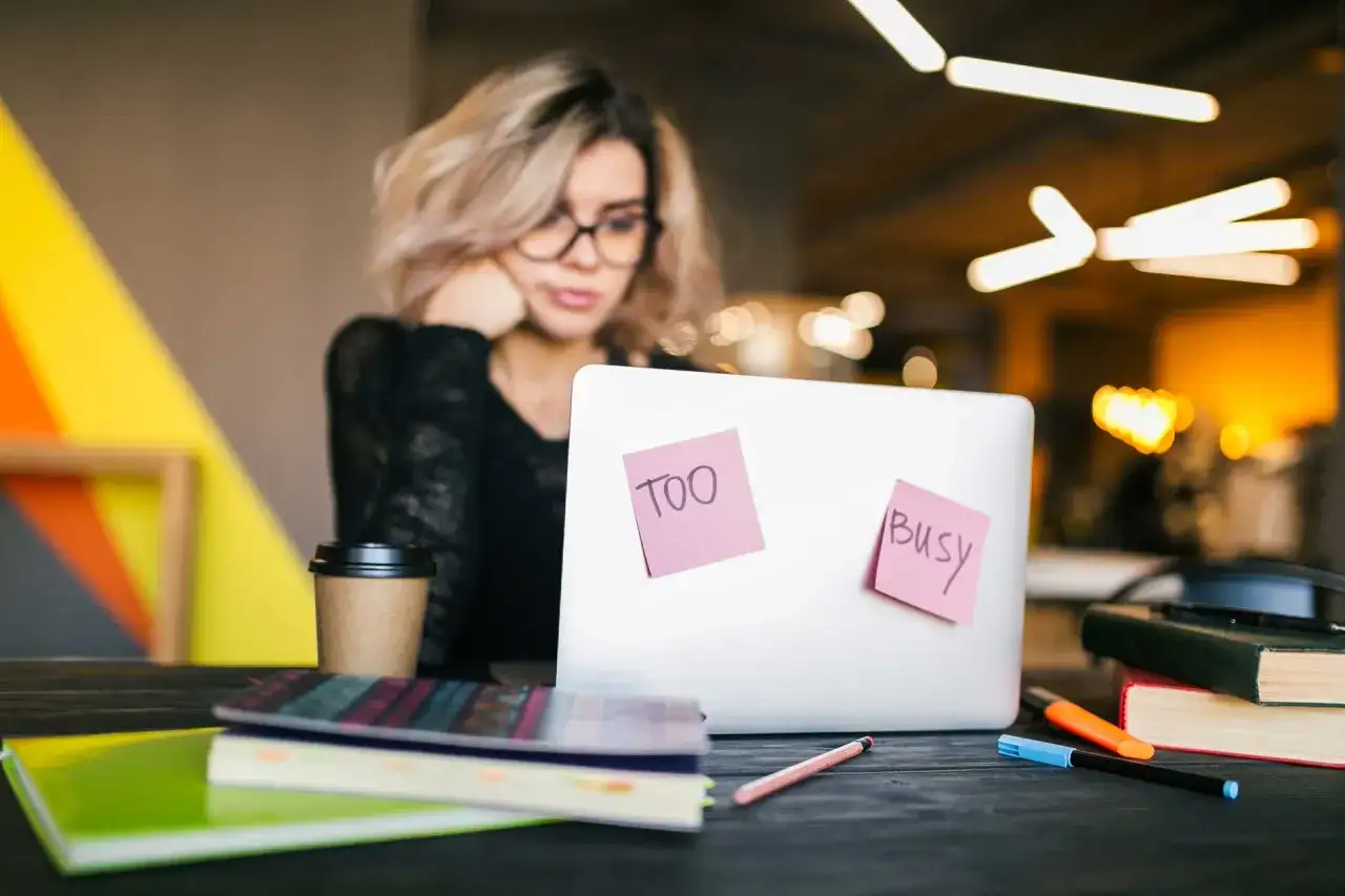 Kobieta z okularami przy laptopie, na którym karteczki "Too busy". Nawet przy natłoku obowiązków, samodzielna nauka angielskiego jest możliwa.
