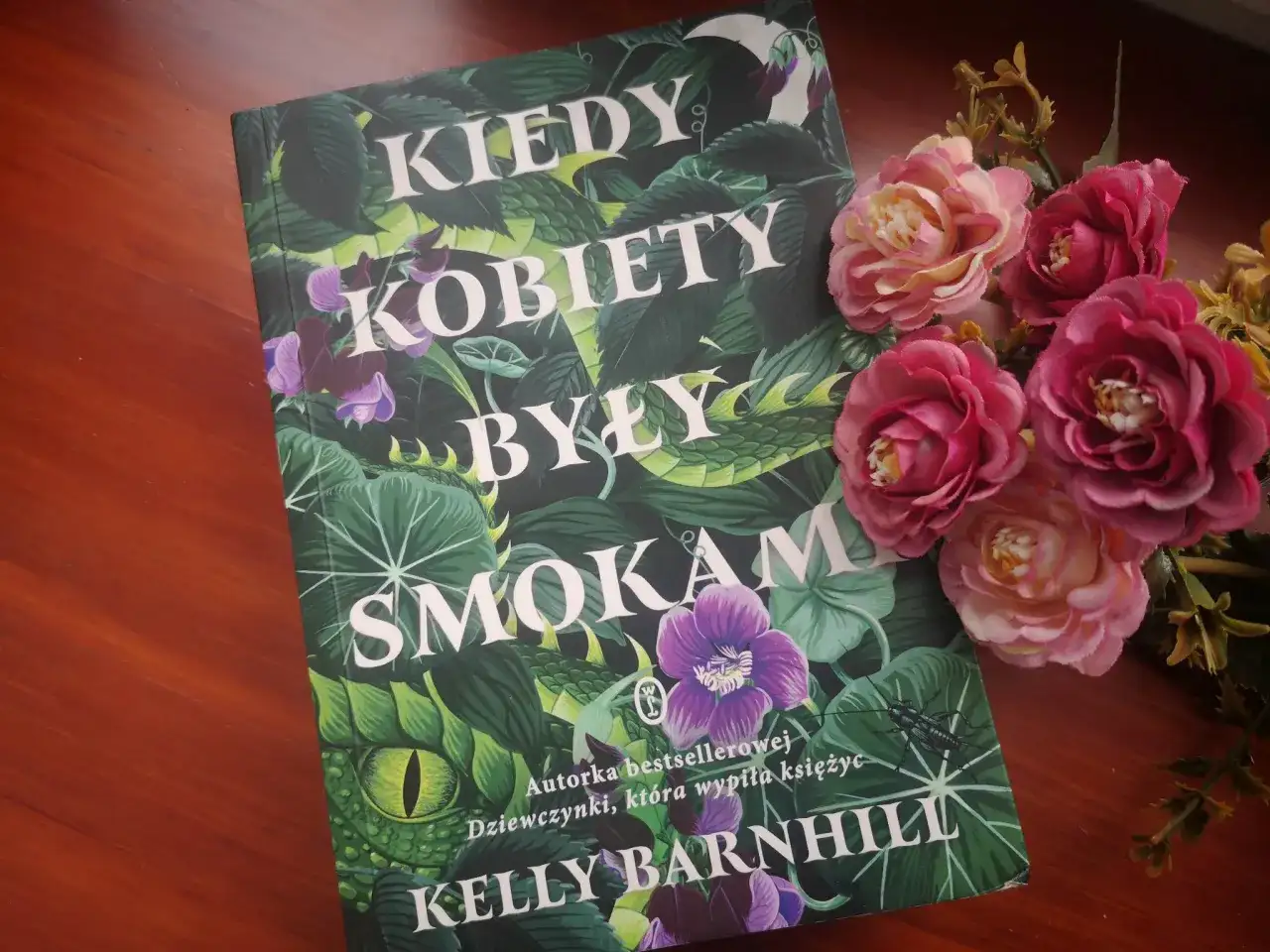 Okładka książki "Kiedy kobiety były smokami" Kelly Barnhill, otoczona bujną zielenią i kwiatami.