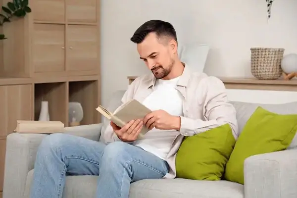 Mężczyzna w jasnej koszuli i jeansach siedzi na kanapie i czyta książkę.